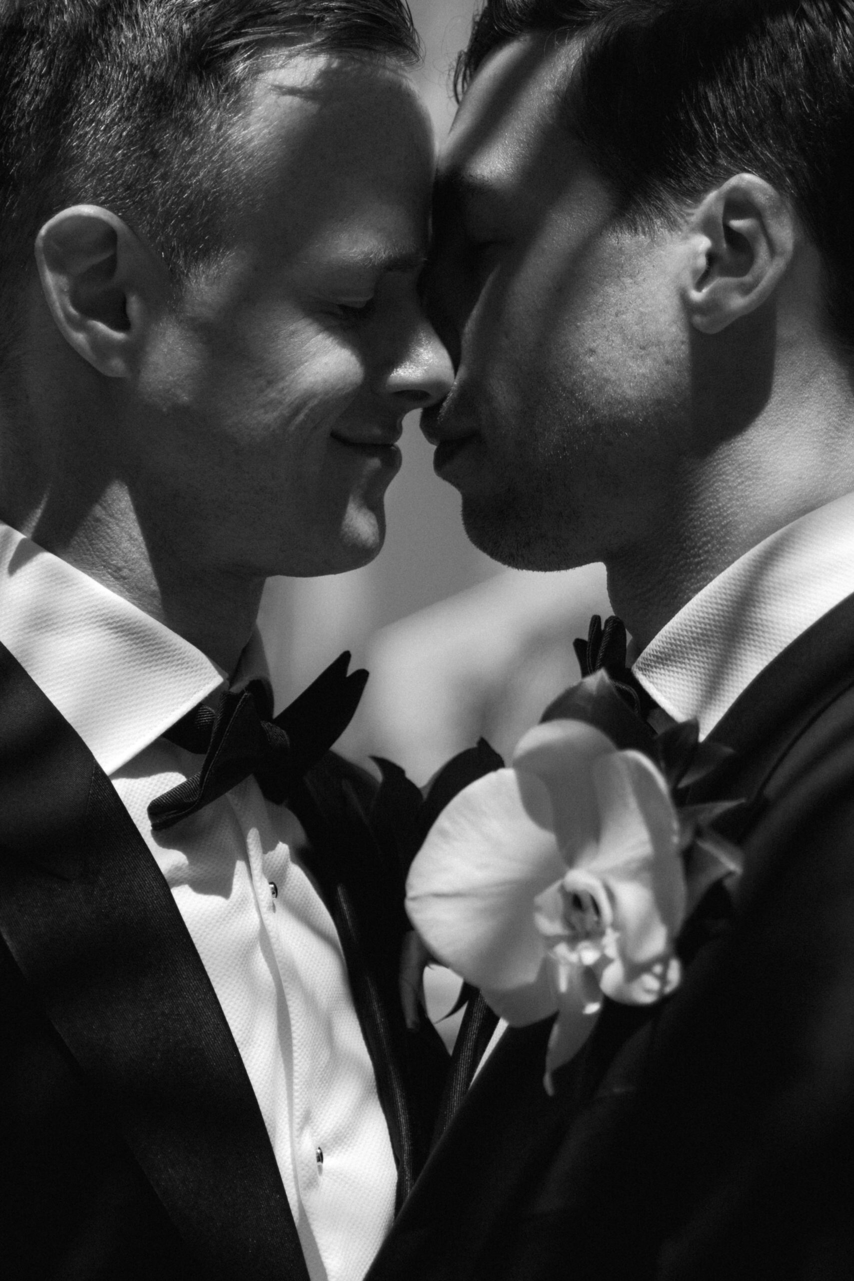 black and white closeup of gay couple embracing in formal tuxedos with palm tree shadows on their faces