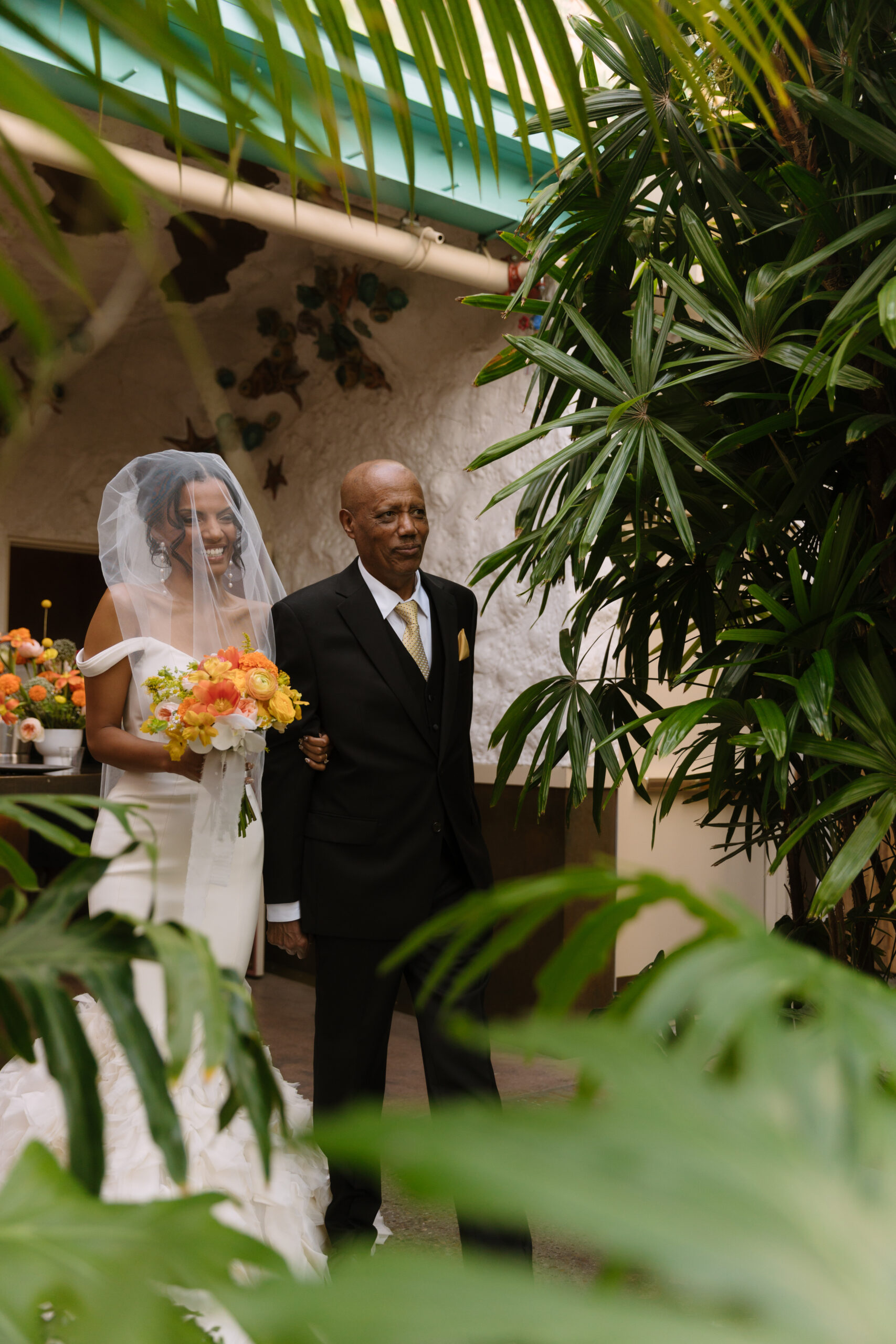 photo shot between tropical plants of bride and her father walking down the aisle at the Grass Room DTLA