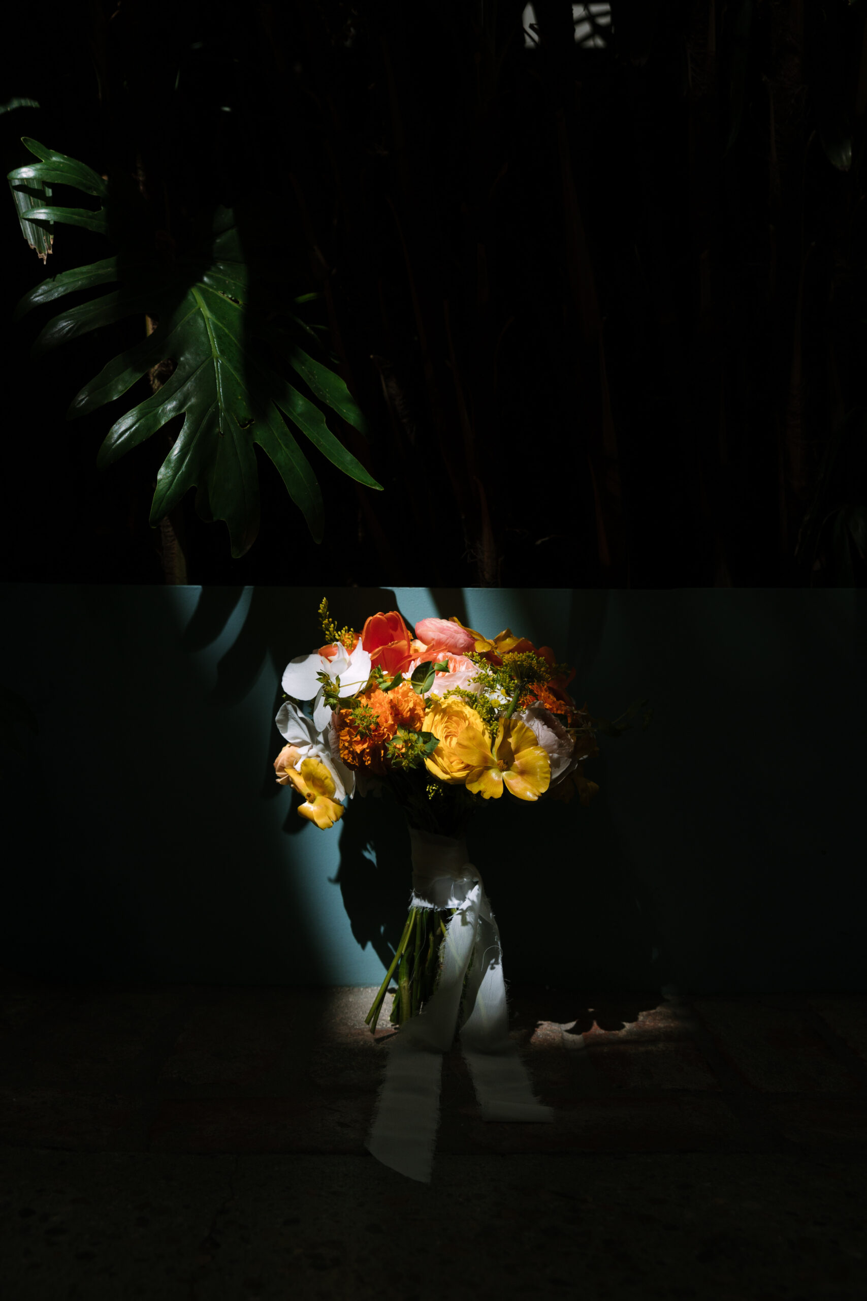 red, yellow, and orange tropical bouquet with white orchids leaning against a turquoise wall under shadow of Monstera plant