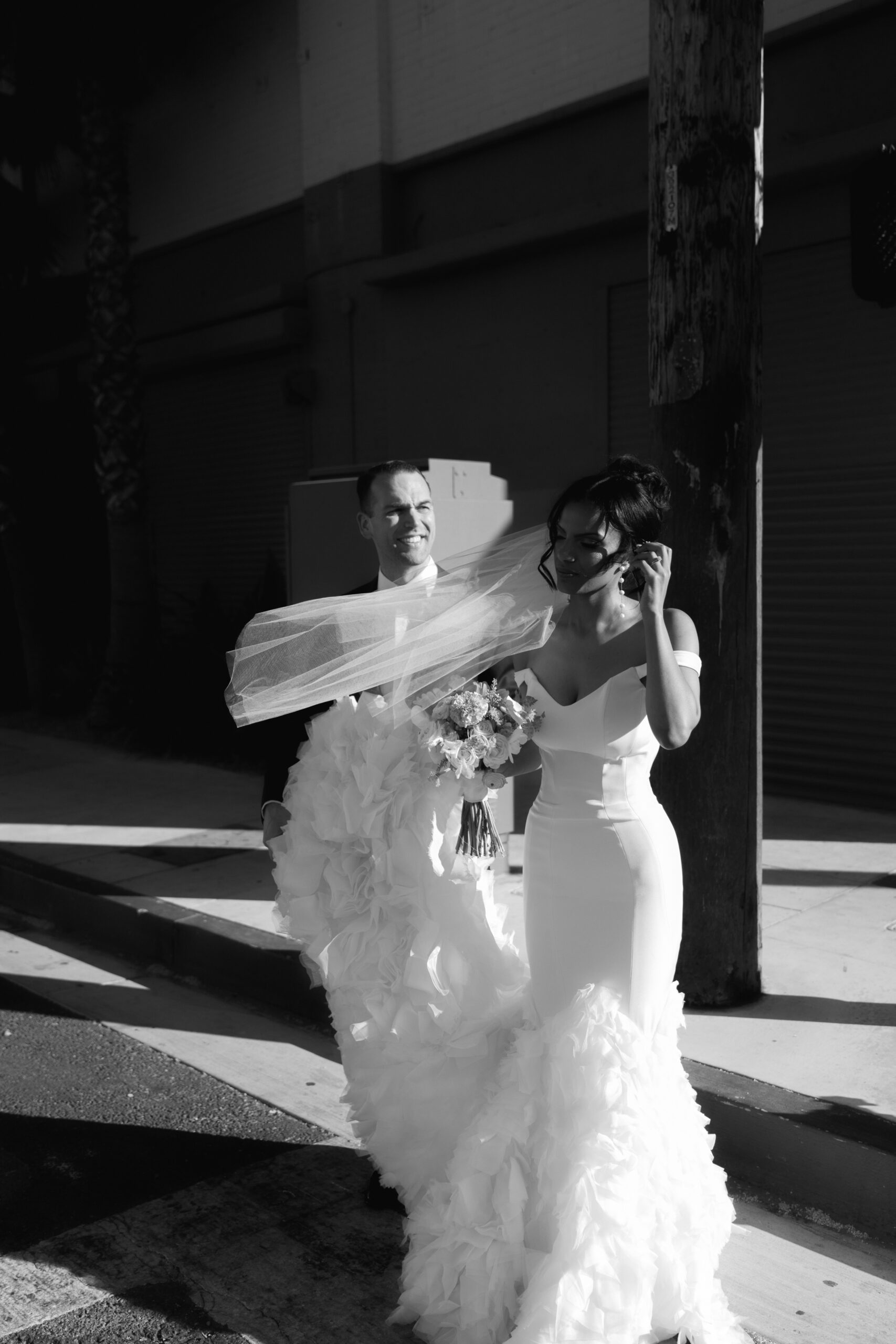 black and white photo of bride adjusting her veil outside in the wind as groom holds the train of her wedding dress