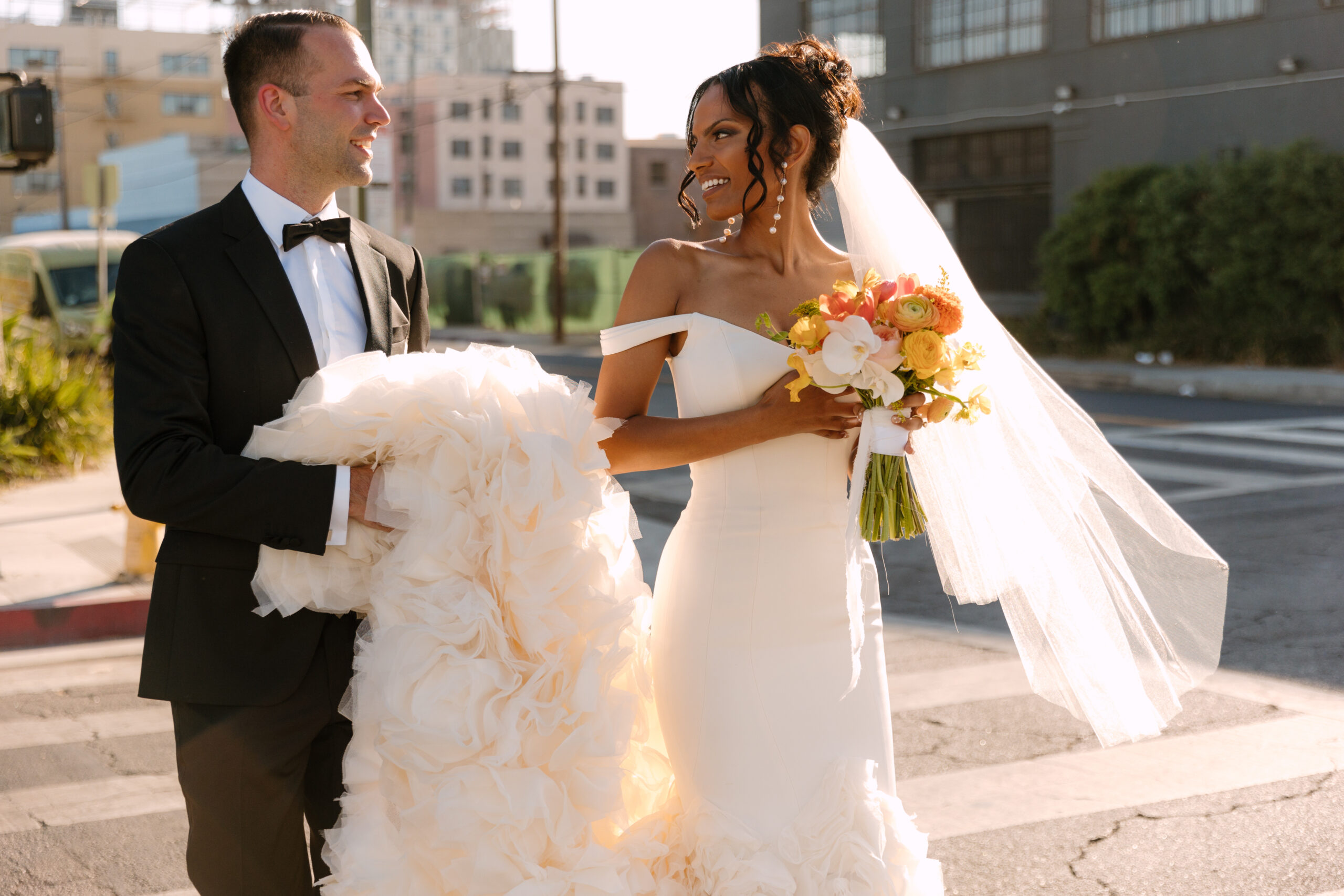 photo of white groom holding Black bride's train as they walk through a crosswalk in DTLA smiling at each other