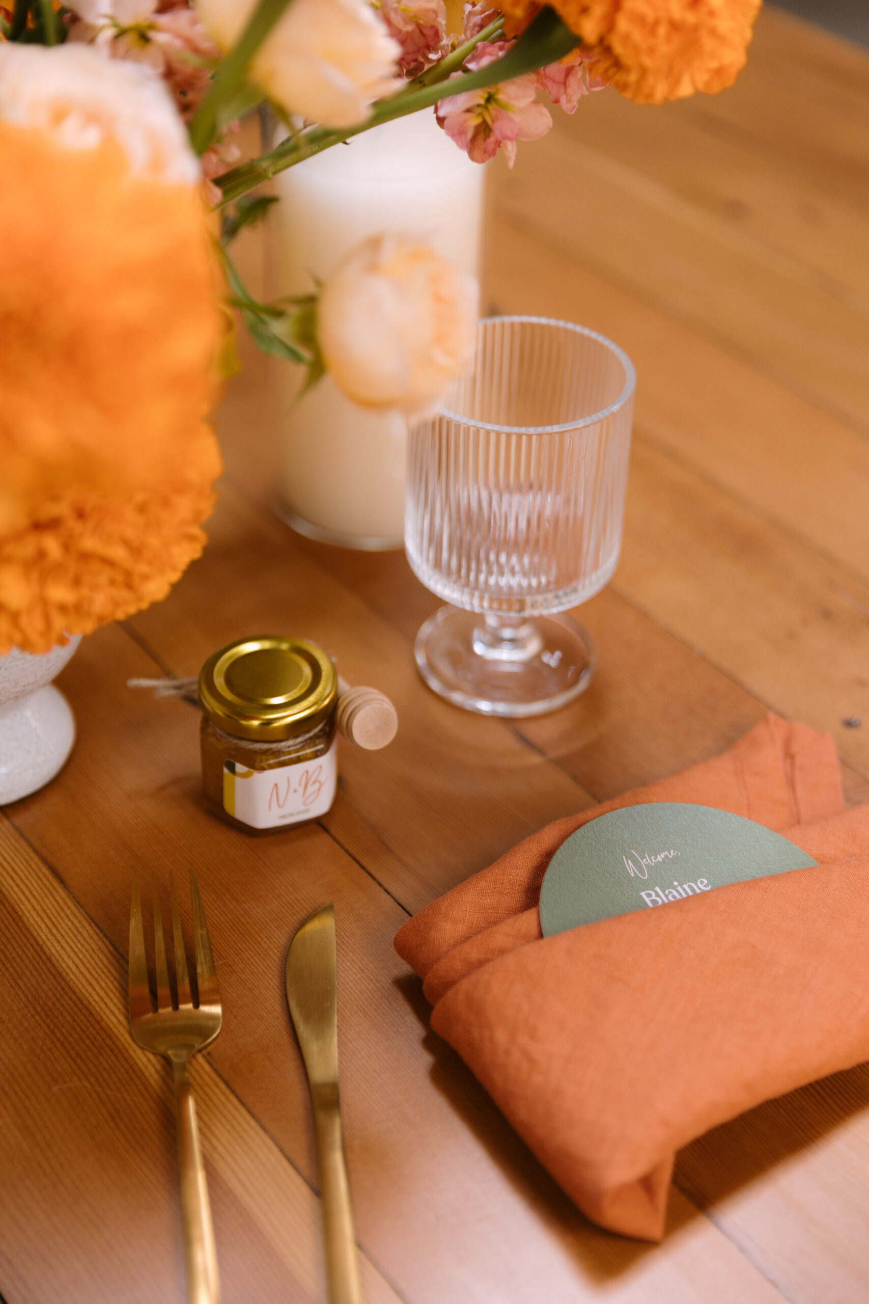 wedding place setting with escort card, fluted water goblet, floral arrangement, gold flatware, and mini bottle of honey