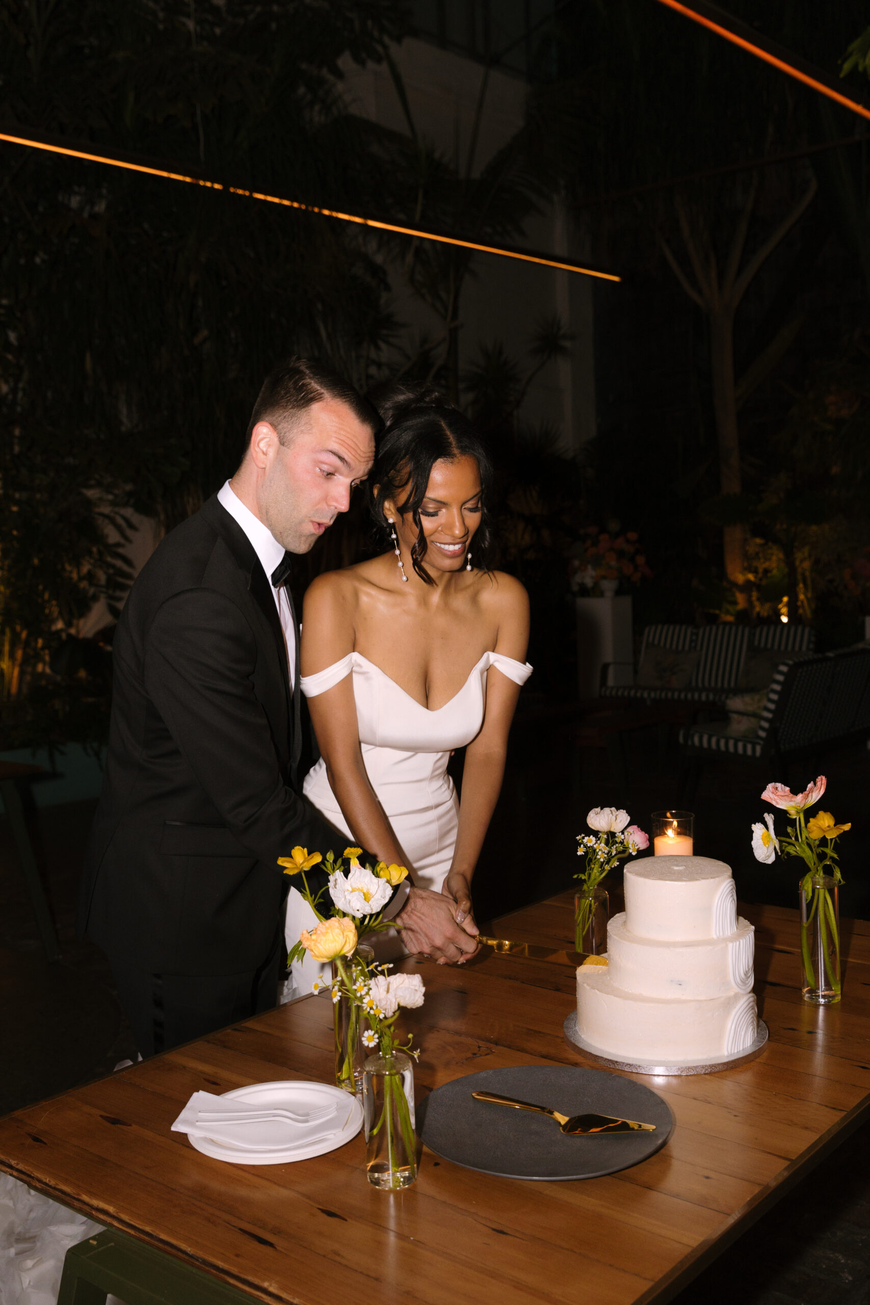 bride and groom cut simple three tier white wedding cake while he makes an excited face