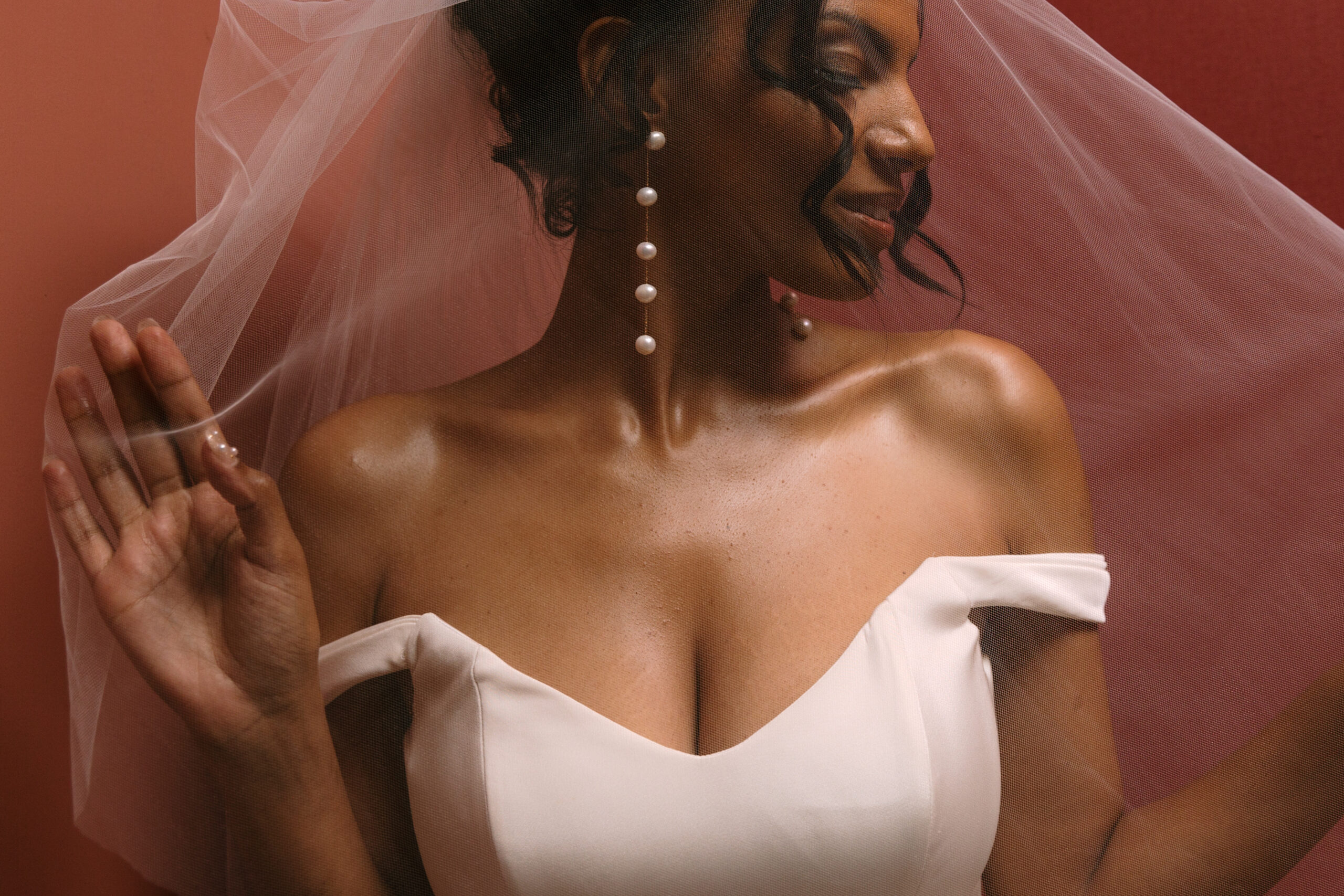 close up of Black bride with long pearl earrings underneath her veil against a red wall