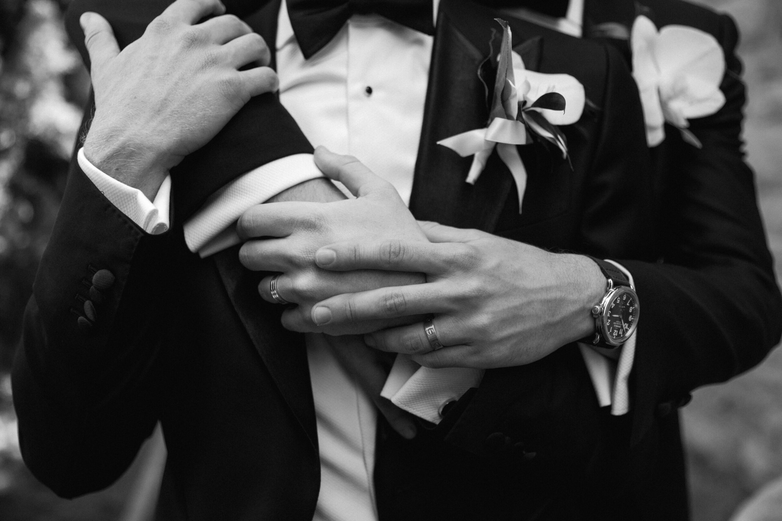 black and white closeup of gay couple back to front embracing their hands and showing wedding bands