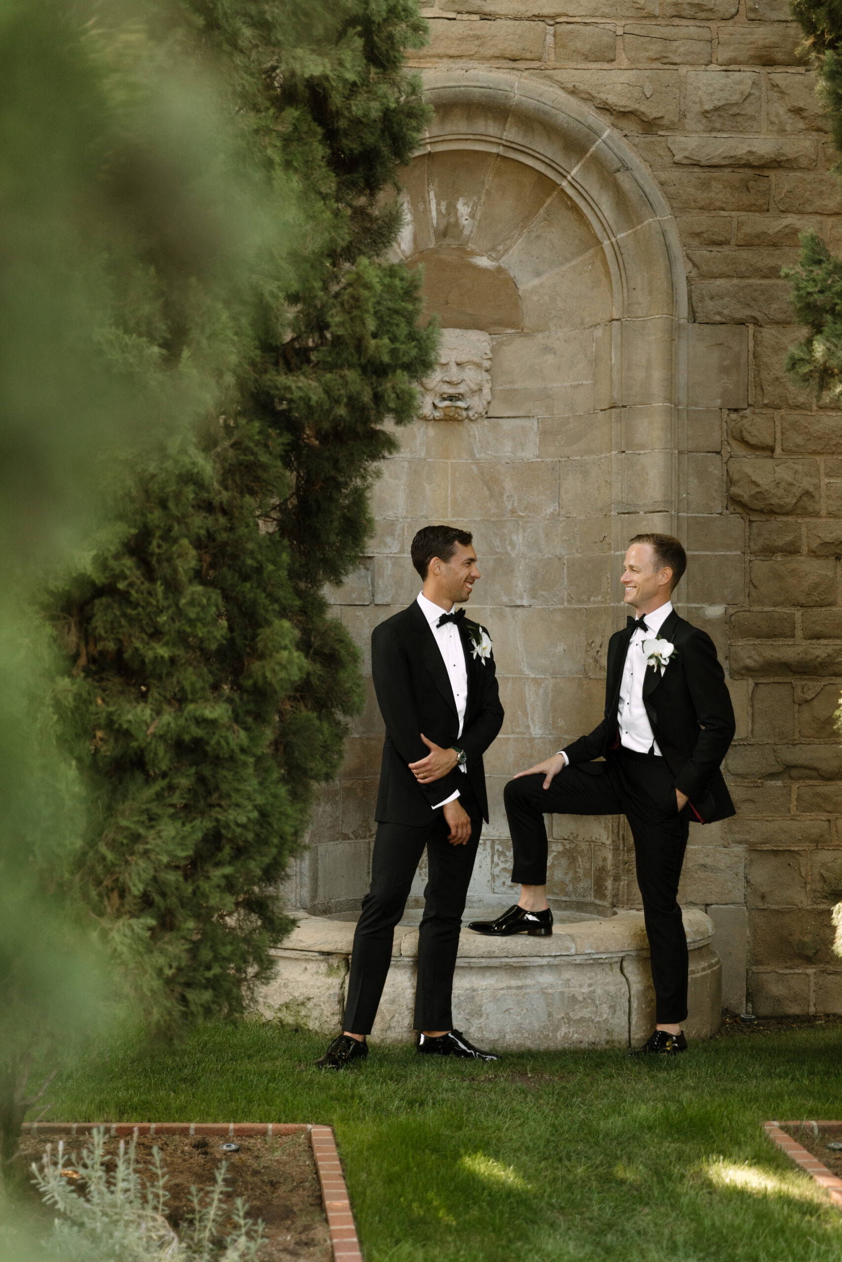 portrait from behind tree of gay couple leaning on fountain at greystone mansion