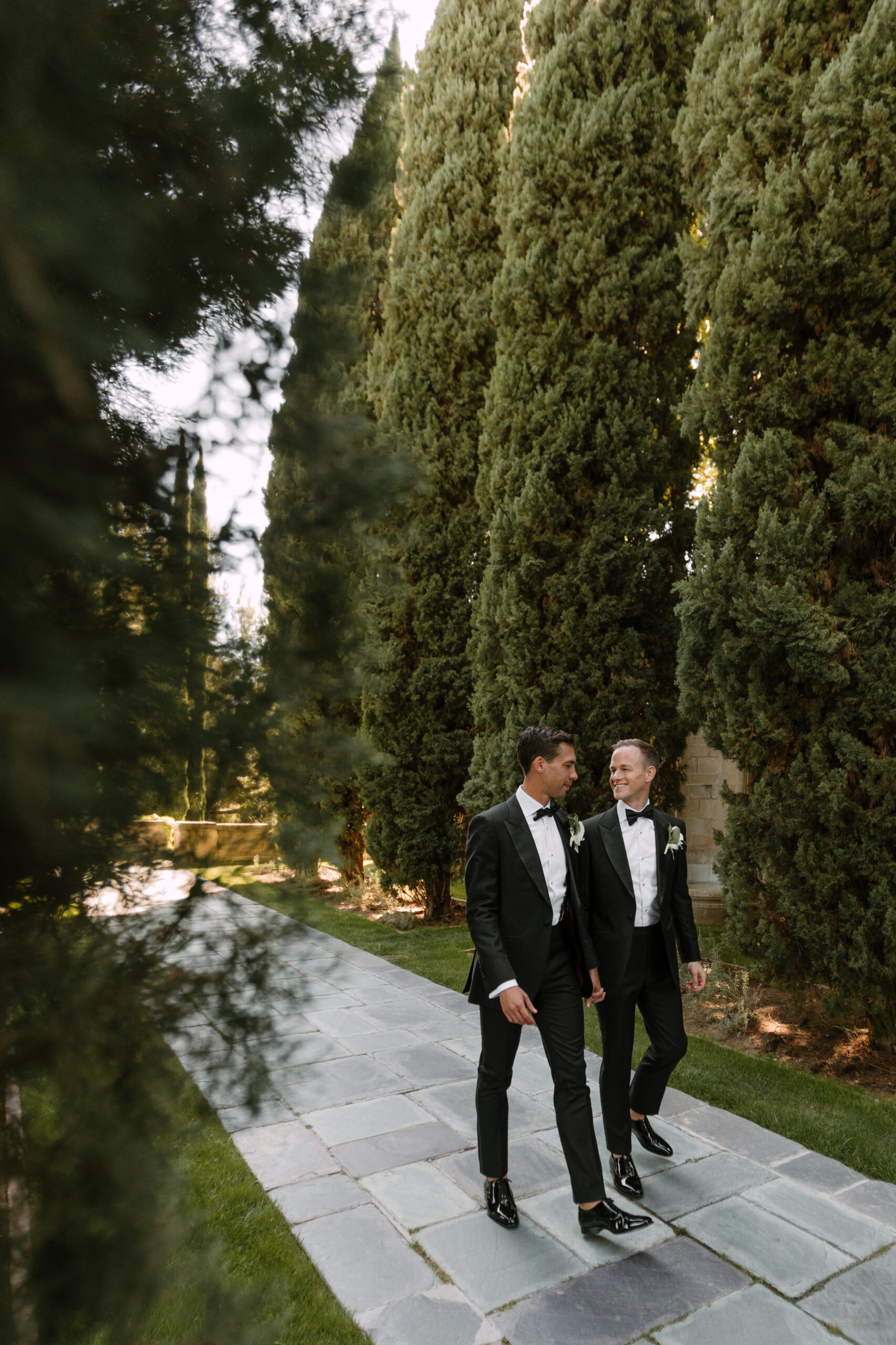 gay couple holding hands looking at each other and walking through Italian Cyprus lined walkway at Greystone Mansion