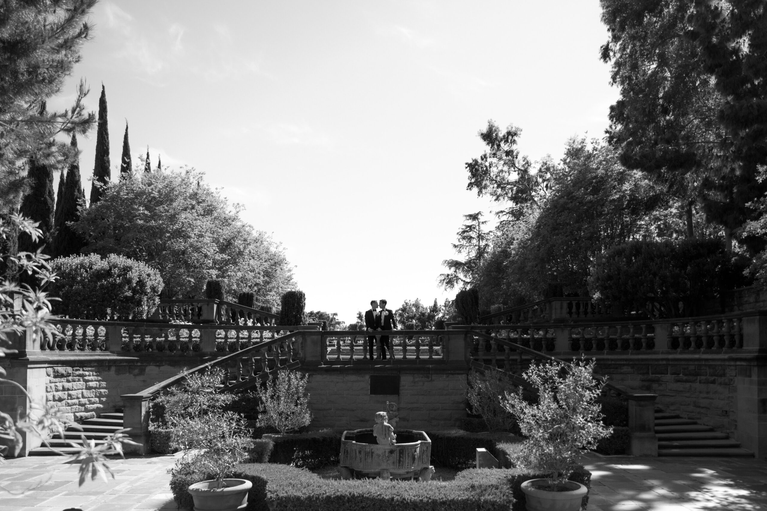 black and white wide portrait of gay couple at the top of garden staircase overlooking Greystone mansion