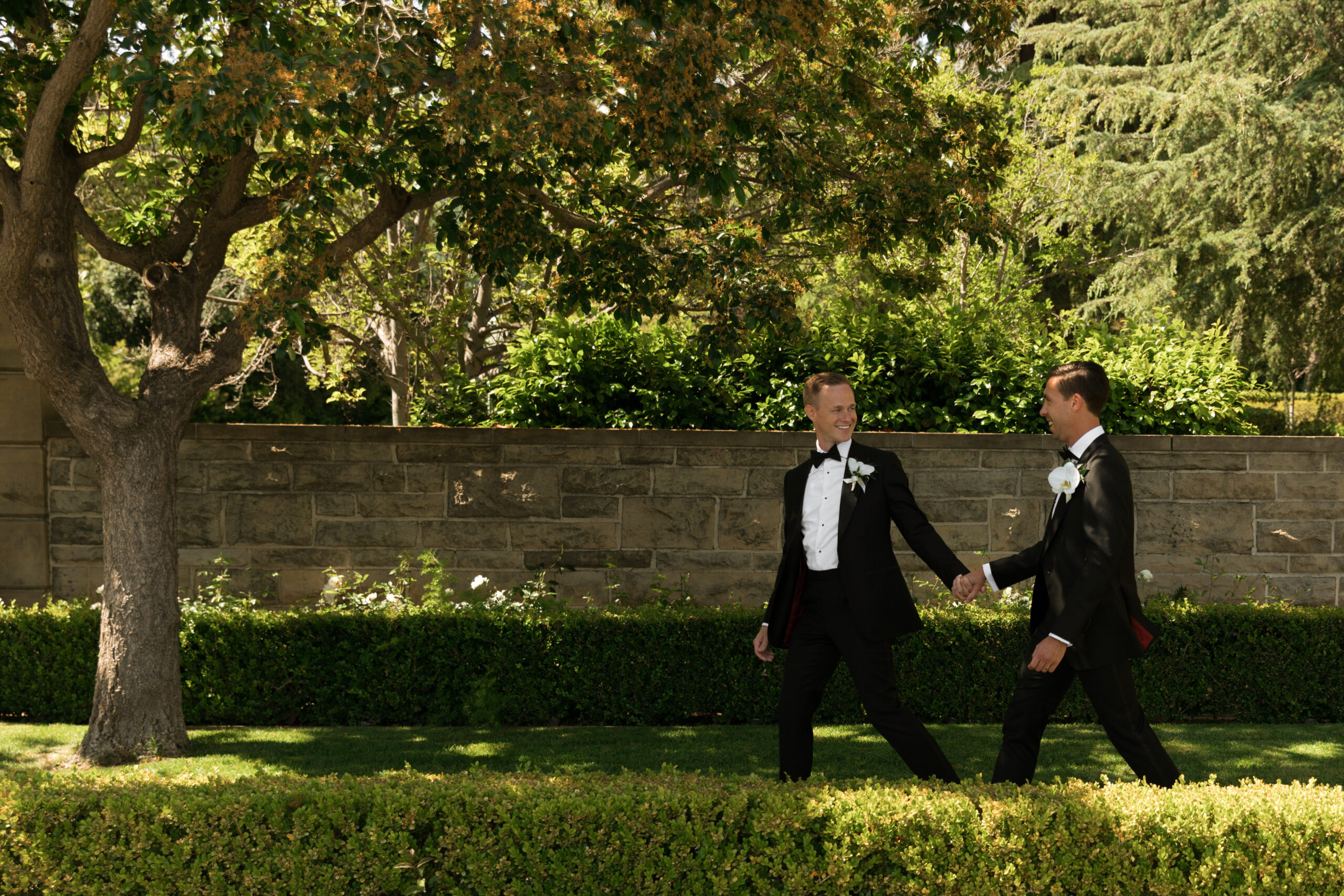 gay couple in formal black tuxes walks through sunny garden at Greystone Mansion