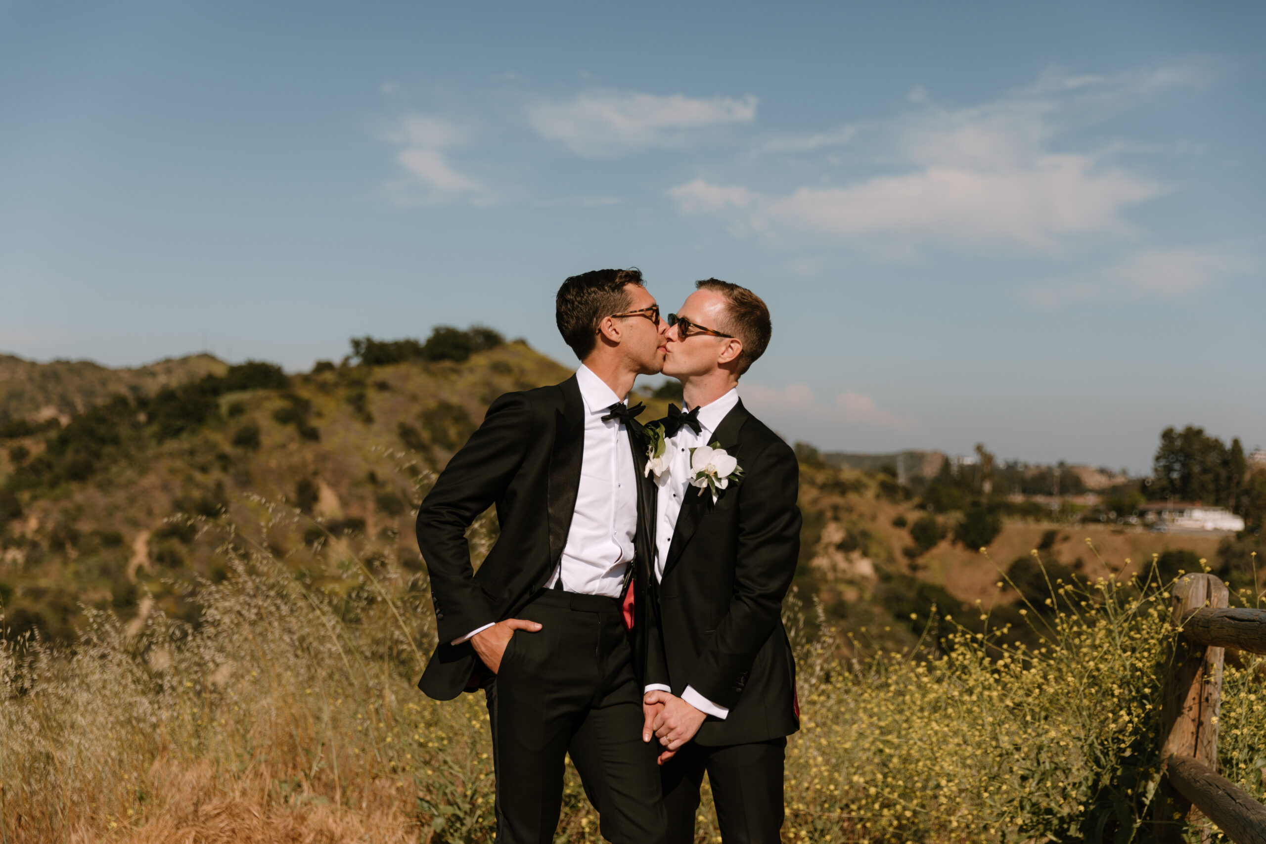 gay couple in tuxedos and sunglasses leans in for kiss overlooking Mulholland Drive