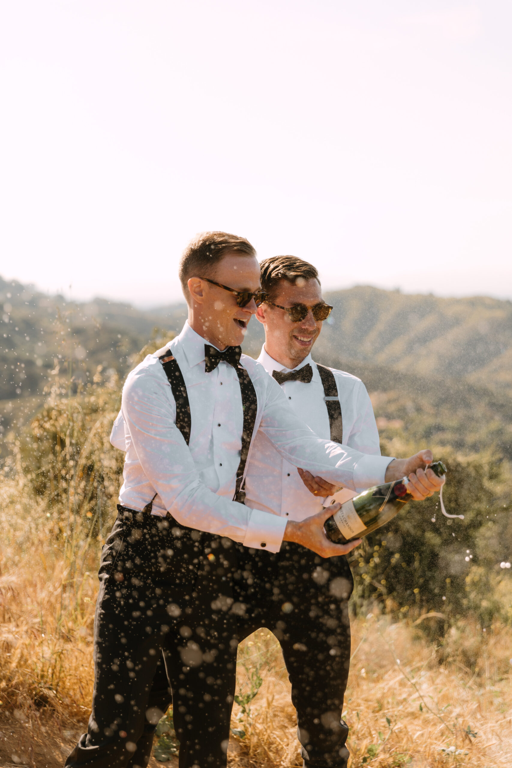 gay couple in suspenders and sunglasses pops foaming champagne bottle overlooking Mulholland Drive