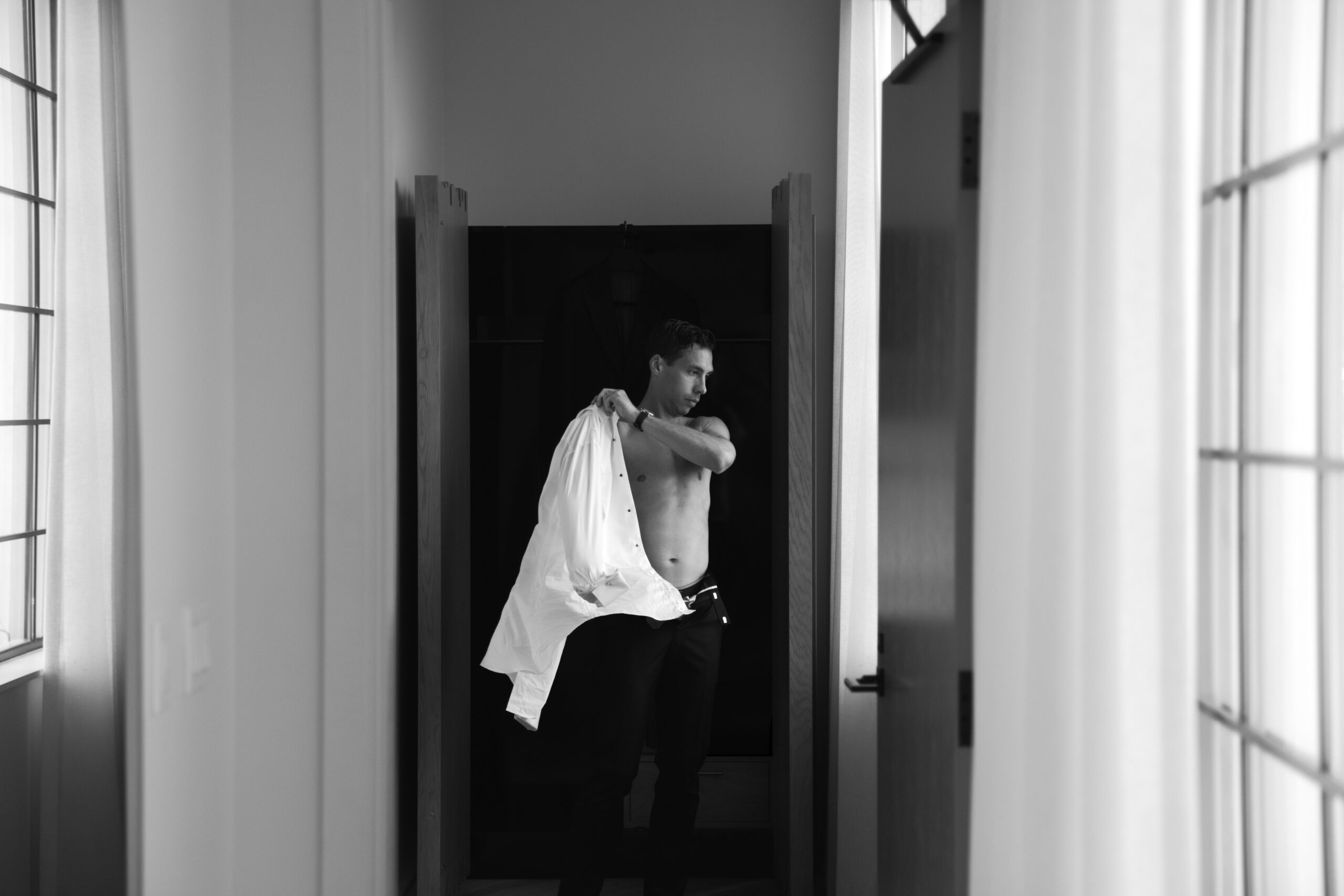 black and white photo of groom in front of window at end of long hallway putting on a white dress shirt