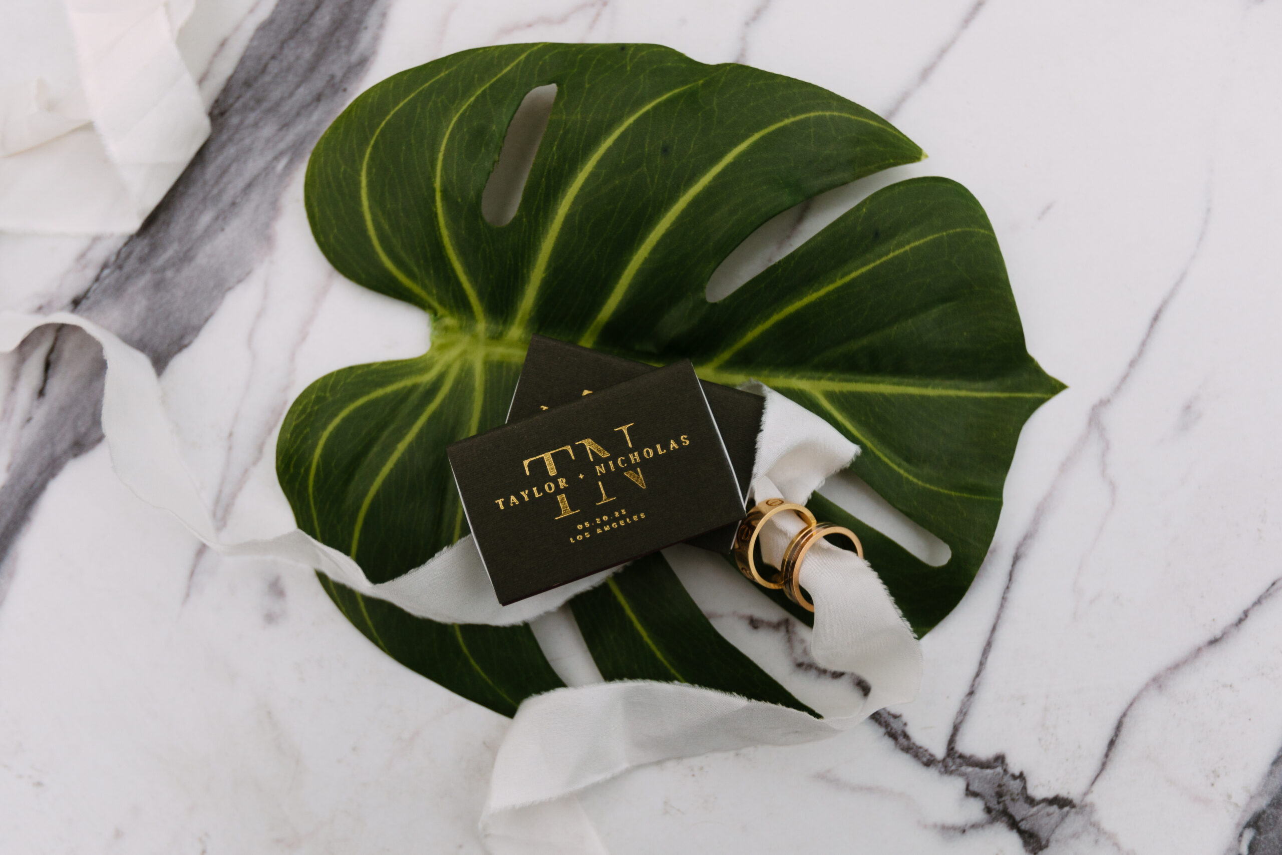flat lay of custom black matchbox, white ribbon, gold Cartier wedding bands, and monstera leaf