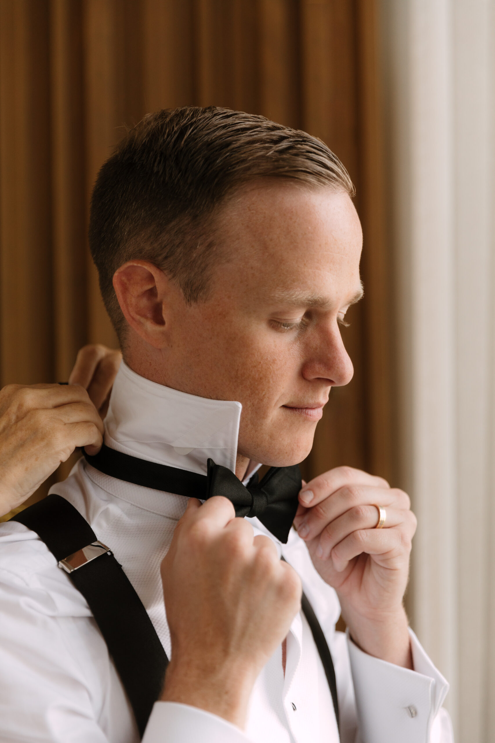 closeup of groom putting on his bowtie with assistance from his mom