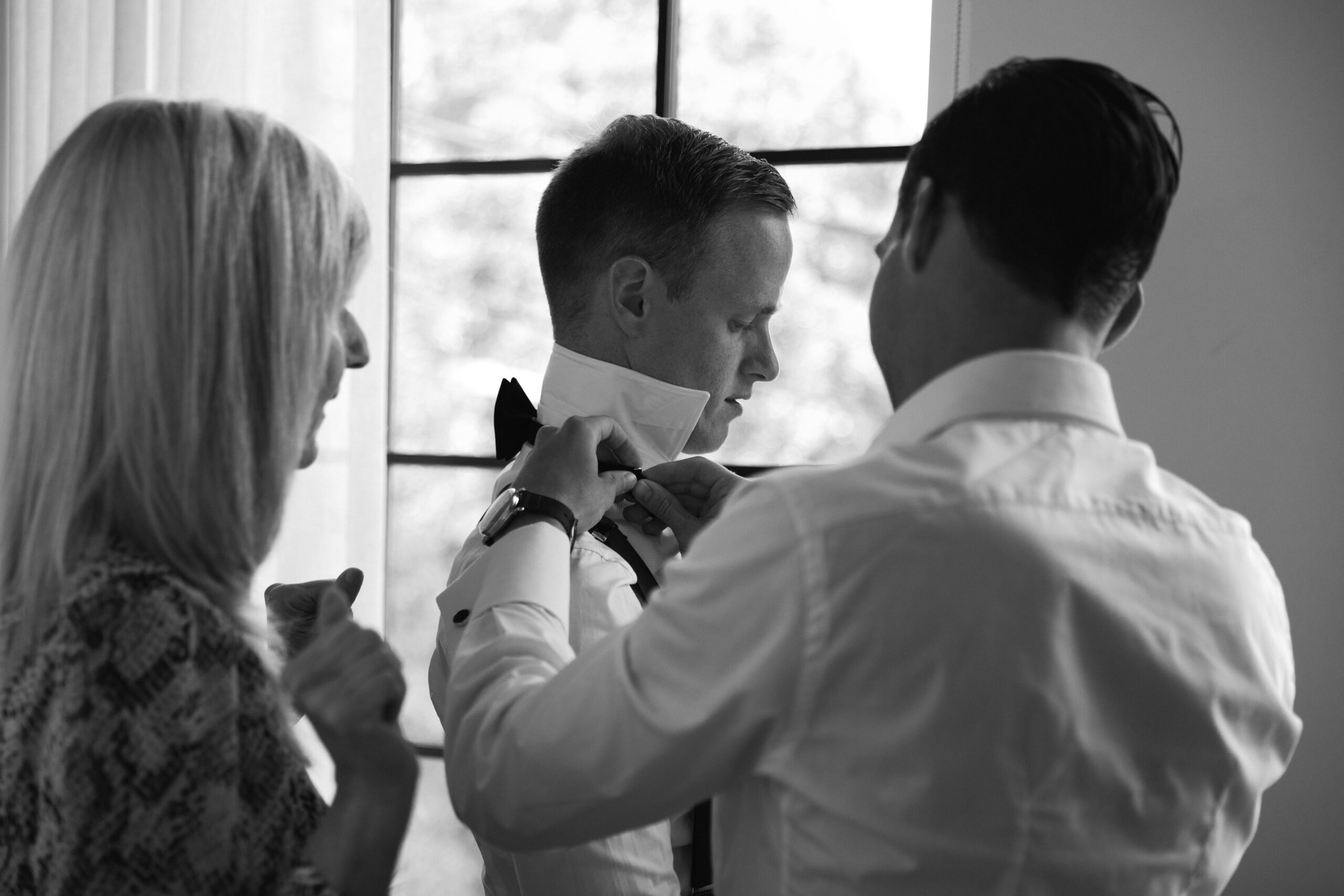 black and white photo backlit by window of gay couple getting dressed for wedding with one mother helping adjust a bowtie