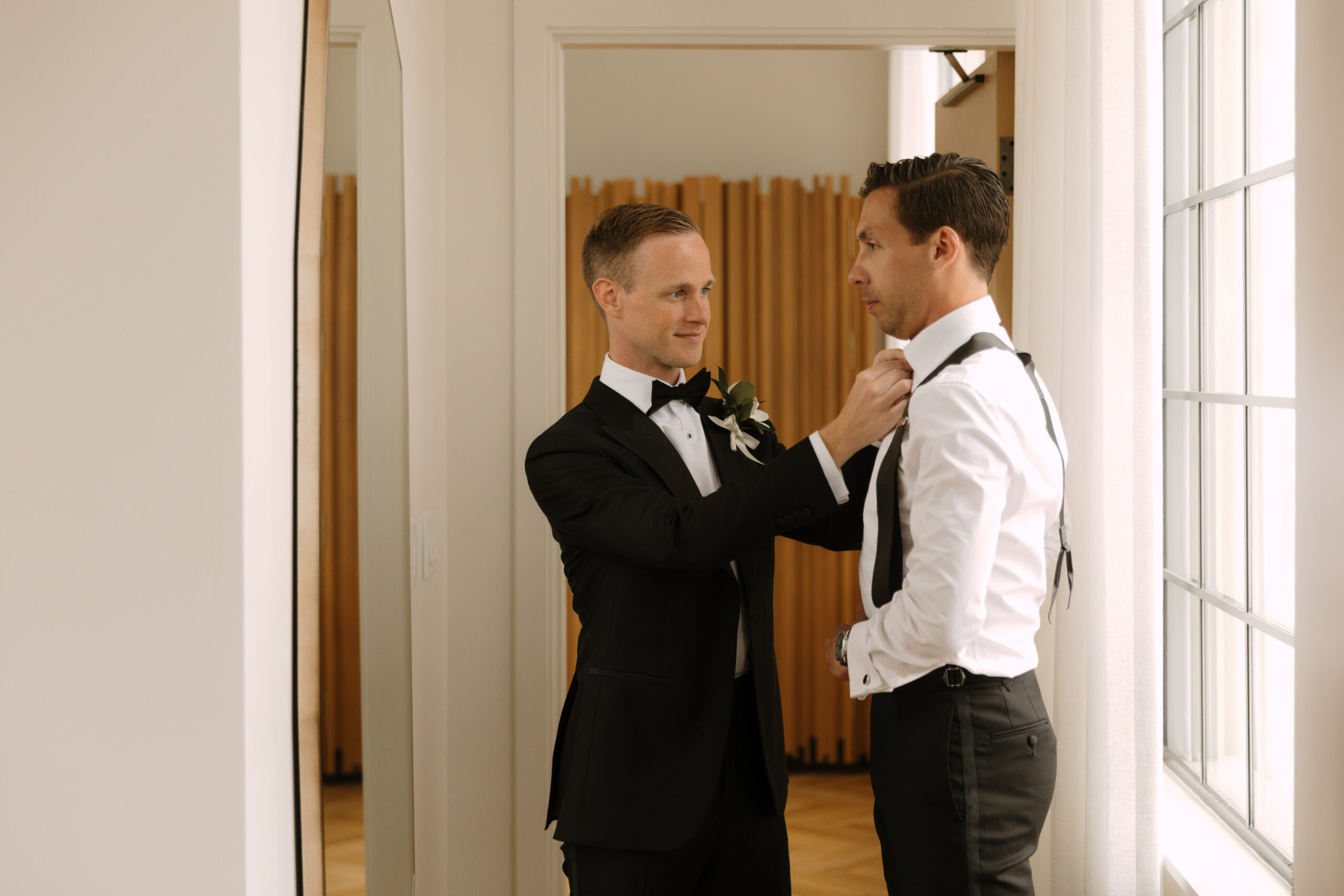 gay groom helps his partner adjust suspenders in front of mirror while getting dressed in Design Suite at Kimpton La Peer Hotel