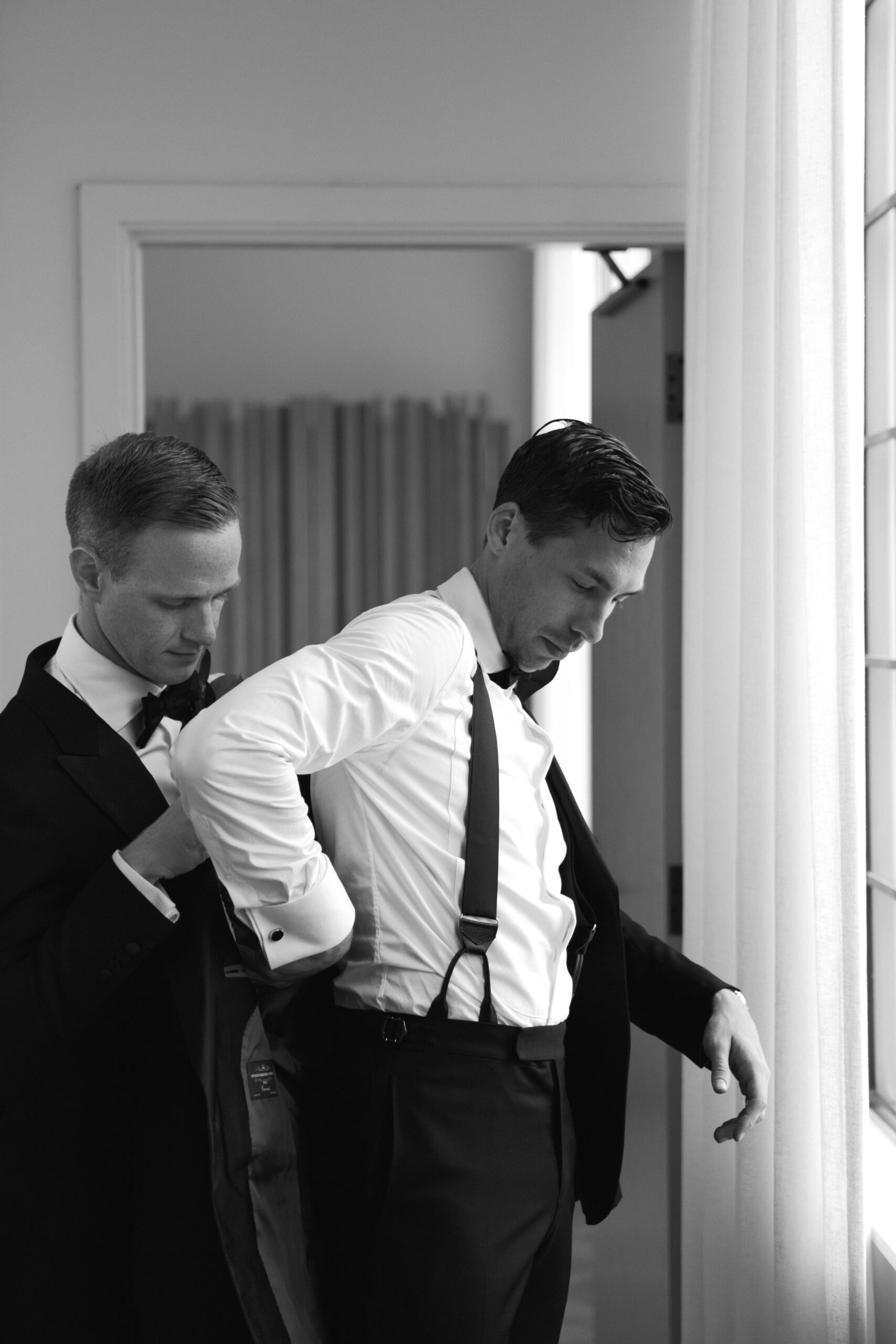 black and white photo of gay groom helping his partner put on his tuxedo jacket