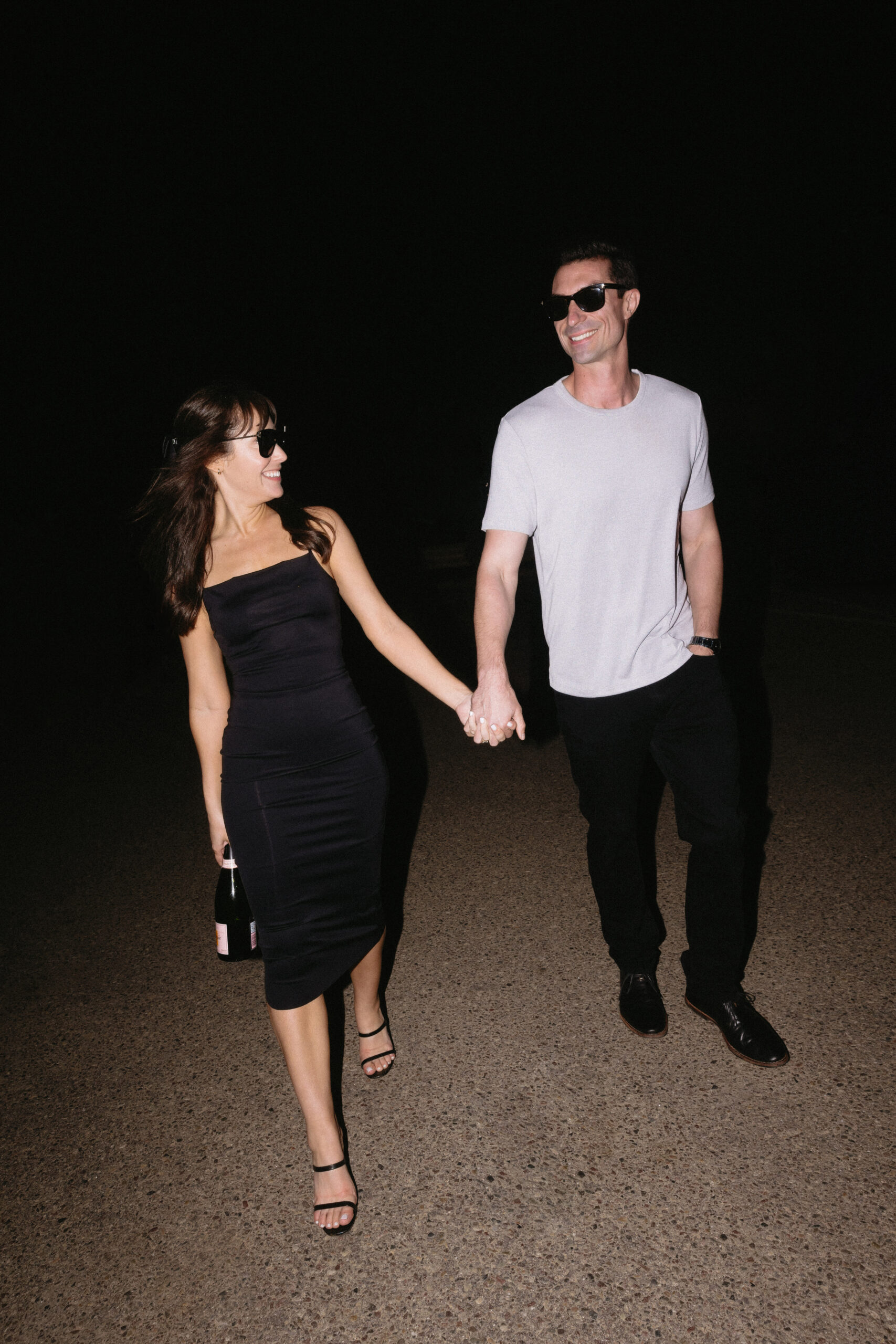 direct flash photo of couple in sunglasses walking hand in hand with a bottle of champagne