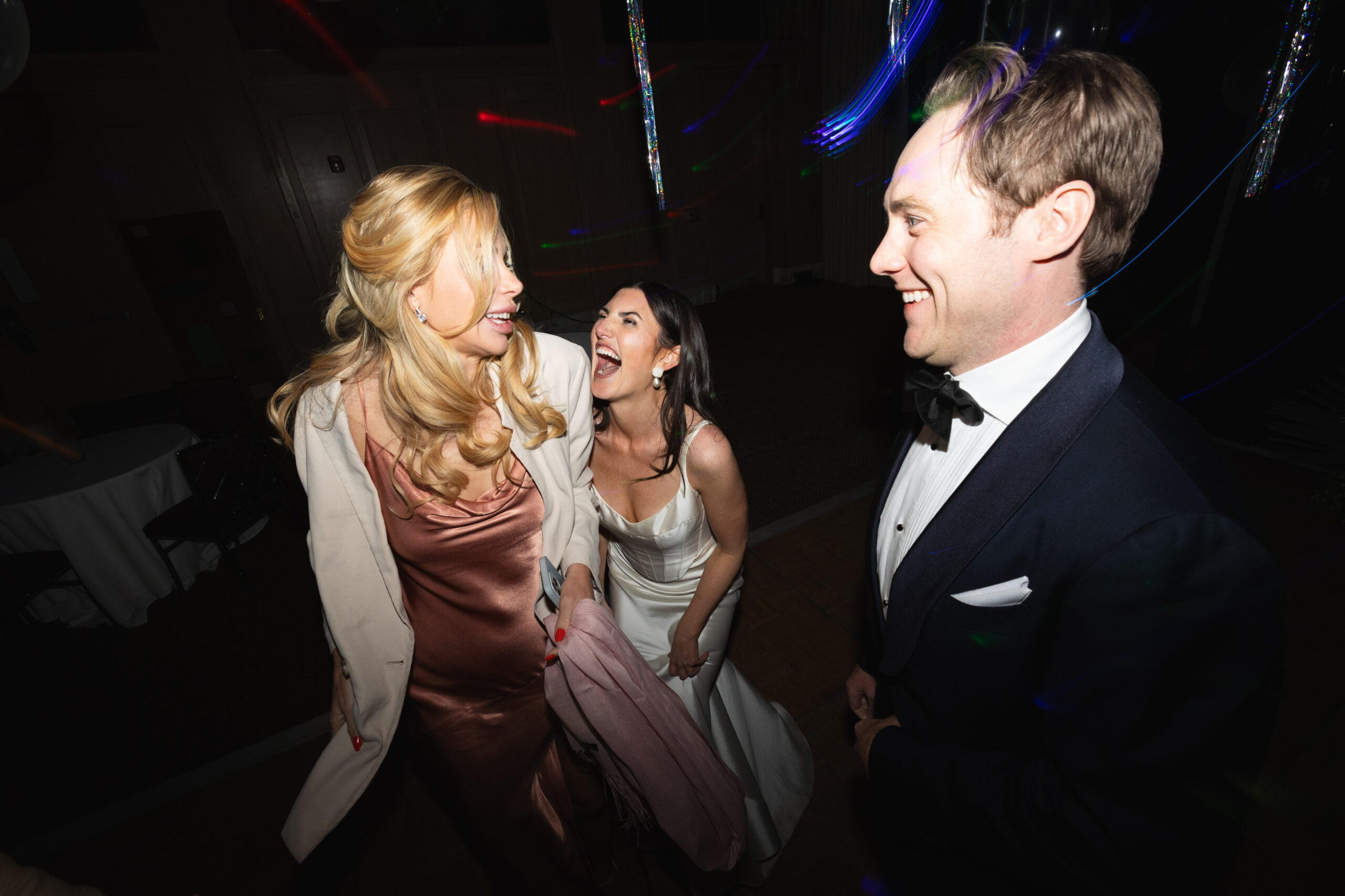 flash photo of bride and groom laughing with guest on dancefloor