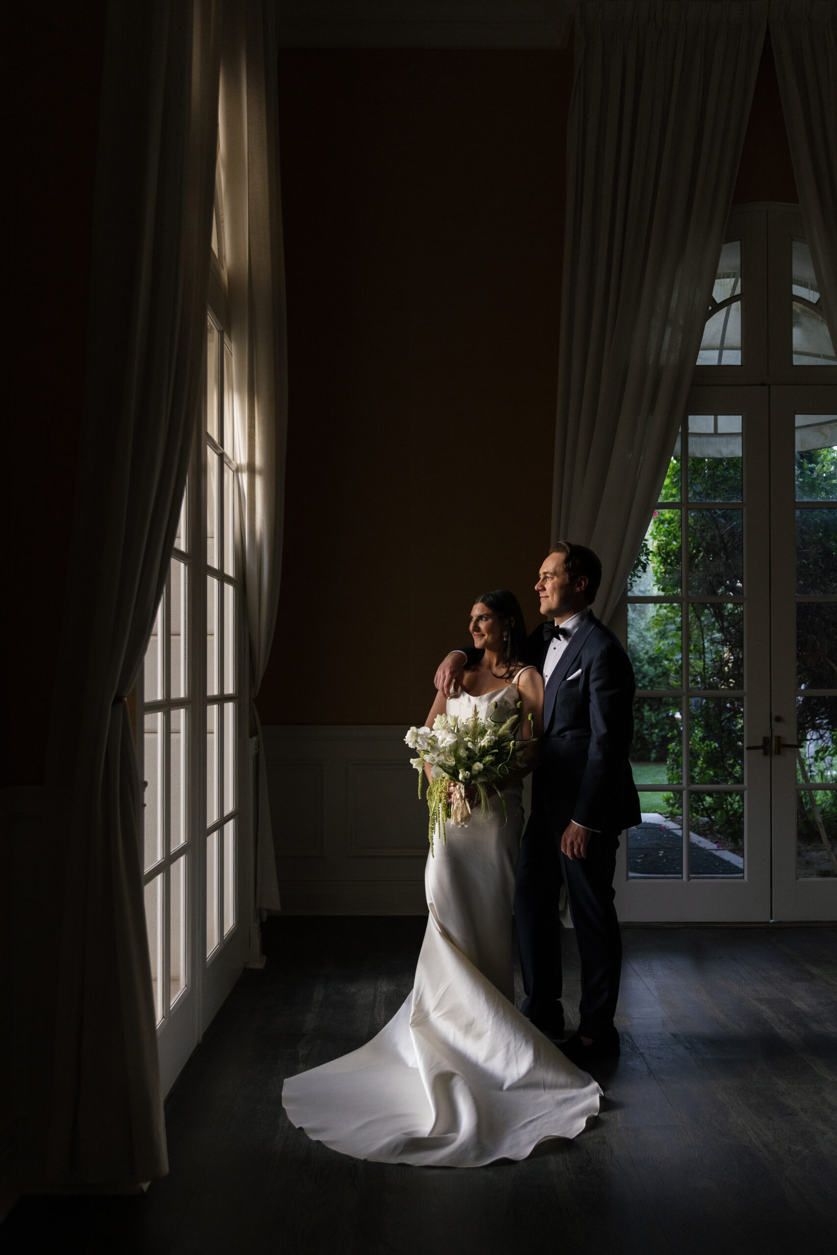 wide shot of groom with arm draped over bride's shoulder in front of windows at Mafia suite at Parker Palm Springs