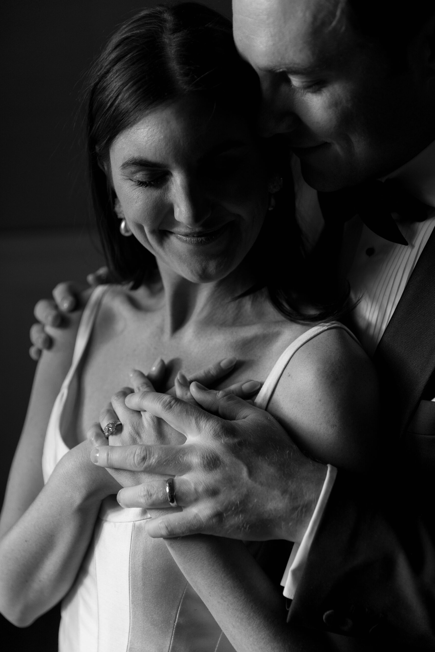 black and white crop of groom's arms around bride showing off wedding rings as she looks down at her shoulder