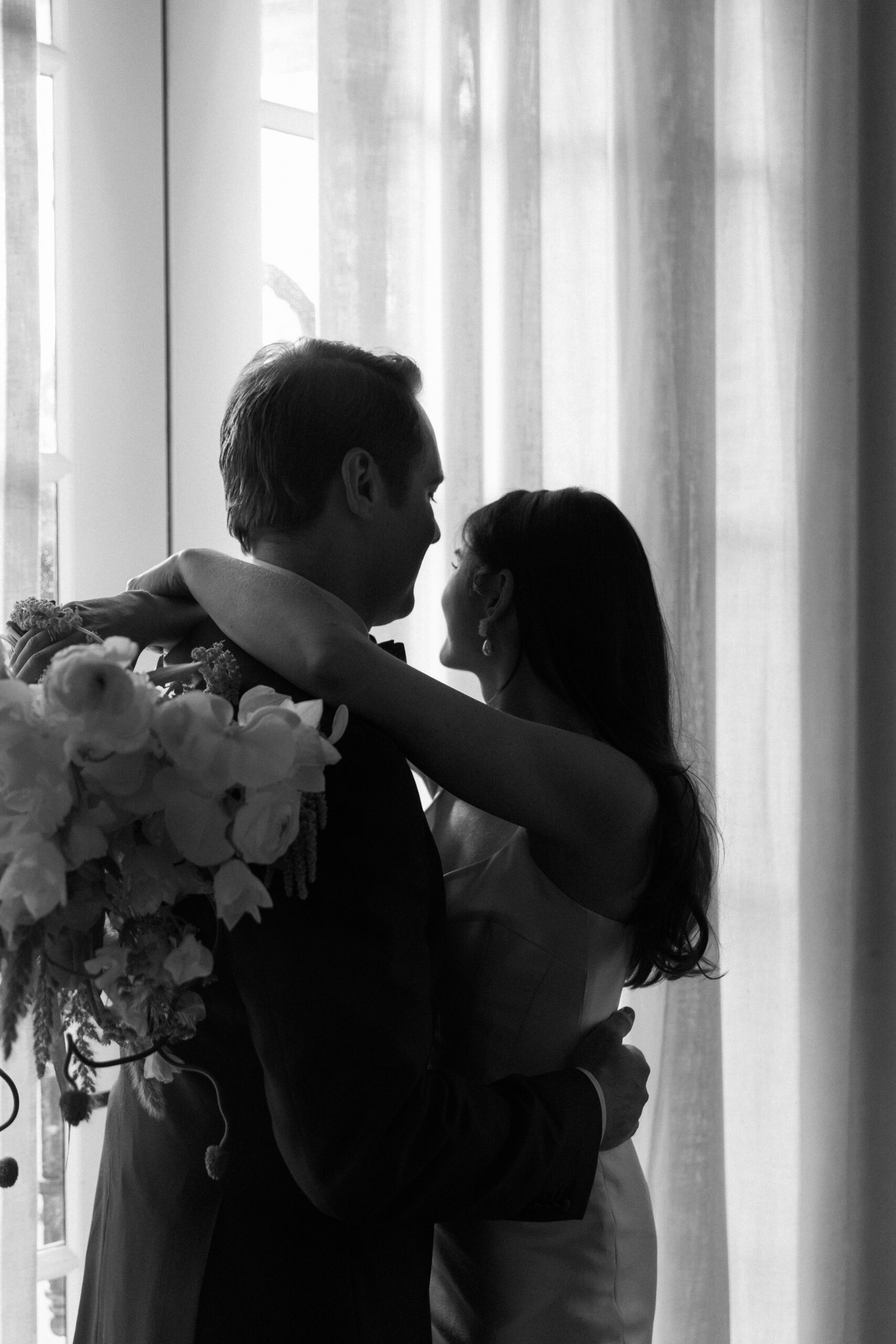 black and white shot from behind of bride and groom with arms around each other in front of window light